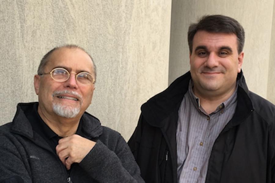 An outside shot of two smiling men in front of a tan, nondescript building. Both are dressed in light black jackets